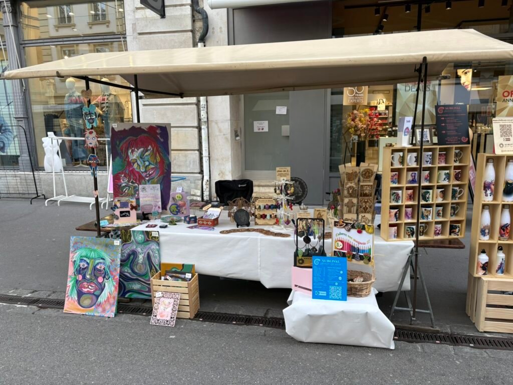 Outdoor market stall with colorful paintings, prints, and crafts displayed on tables and on a wooden shelf outside a shop under a beige canopy.