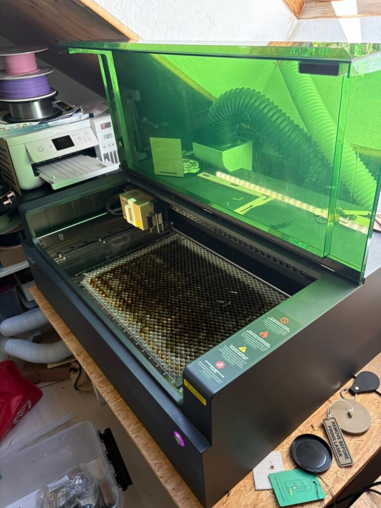 Green-tinted laser cutter on a workbench with the lid open and a honeycomb cutting bed visible; printer and spools nearby in a workshop.